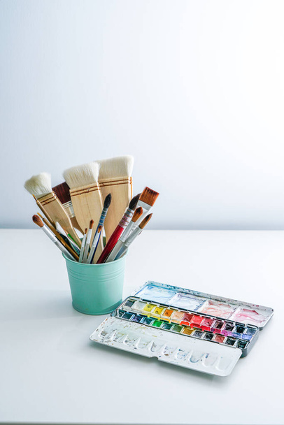 Watercolor palette and picture brushes on white table. Watercolor painting tools house on white desktop - Photo, Image