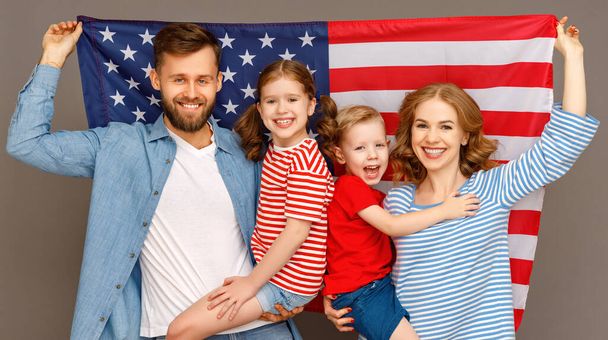 Boldog szülők amerikai zászlóval és vidám gyerekek ölelik egymást, és a kamerába néznek, miközben a Függetlenség Napját ünneplik a szürke backgroun ellen. - Fotó, kép