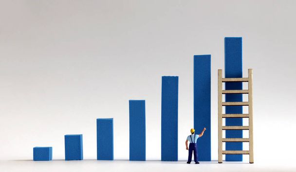 A miniature man points to a blue bar graph with a mini wooden ladder. - Фото, изображение