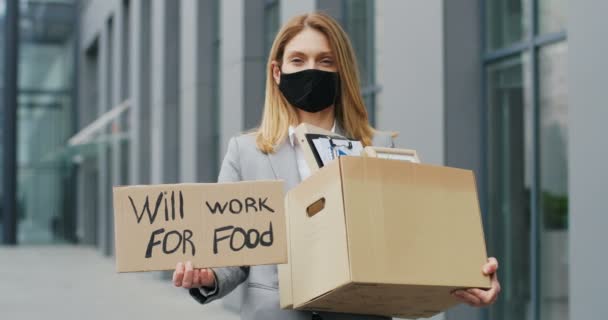 Porträt einer kaukasischen arbeitslosen Frau mit Maske, die draußen steht und ein Plakat mit den Worten Will work for food zeigt. Gefeuerte weibliche Arbeitslose und Kiste mit Bürokram. - Filmmaterial, Video