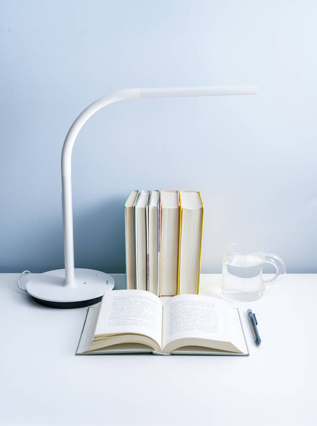 Un libro abierto se coloca sobre una mesa blanca con una lámpara. Una pila de libros cuidadosamente arreglados y un vidrio transparente sobre una mesa blanca
 - Foto, Imagen