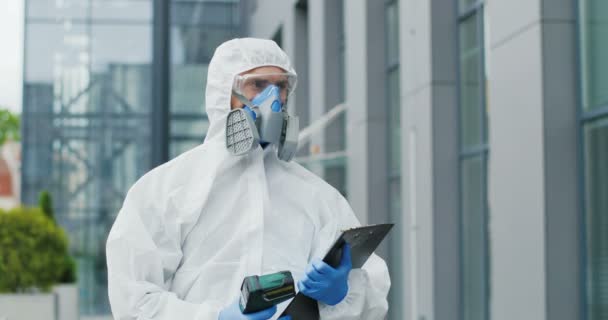 Caucasian man in white suit, goggles, gloves and respirator walking street in town and check. Male healthcare worker of disinfection service holding folder and contactless thermometer outdoor Pandemic - Πλάνα, βίντεο