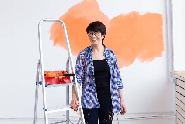 Pretty smiling middle-aged woman painting interior wall of home with paint roller. Redecoration, renovation, apartment repair and refreshment concept. - Photo, Image