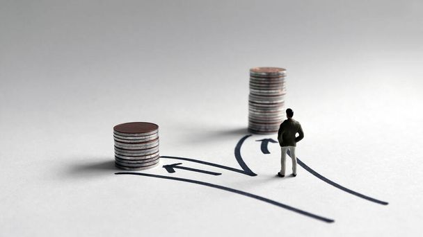 Ένας άνθρωπος μινιατούρα στέκεται μπροστά από δύο μονοπάτια με μια στοίβα από κέρματα διαφόρων υψών. - Φωτογραφία, εικόνα