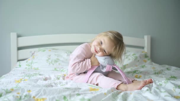 portret. klein meisje in pyjama spelen met een speelgoed konijn op het bed. - Video