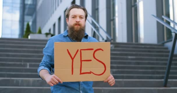 Portrait shot of Caucasian male activist with beard holding poster Yes at demonstration or protest. Single protest outdoor. Activism concept. Man showing table with word yes. - Footage, Video