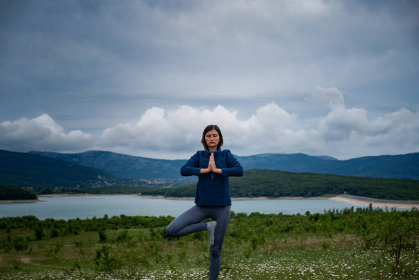 Arazide yoga yapan genç bir kadın yoga ağacı pozu ya da asana Vrikshasana tre pozu veriyor. Doğayla uyum, sağlık ve zindelik kavramı. Kendini analiz ve vicdan muhasebesi. - Fotoğraf, Görsel