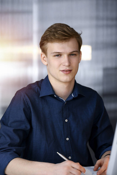 Young blond businessman working with computer in in a darkened office, glare of light on the background - Foto, afbeelding