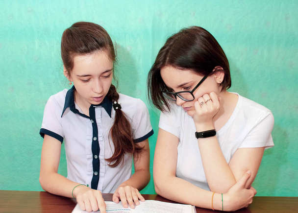 Two Schoolgirls at the table with a book, one explaining something to the other. Medium plan, turquoise background. - Photo, image