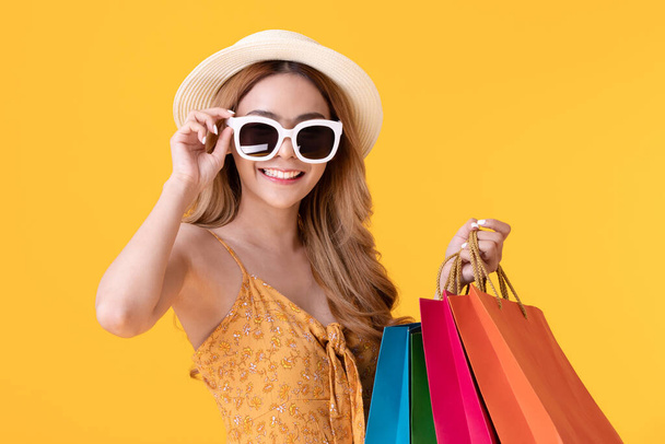 Happy young asian woman wearing hat and Sunglasses holding shopping bags smile and cheerful in summer shopping sale,isolated on yellow background - Фото, изображение
