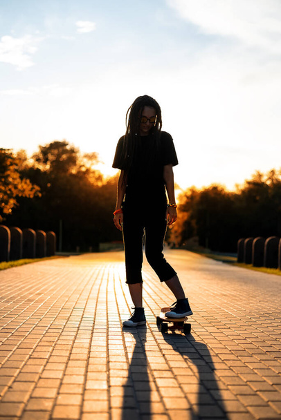 girl in sunglasses stands on a skateboard in the rays of a beautiful sunset. silhouette of a hippie with dreadlocks on a longboard in a park - Foto, Bild