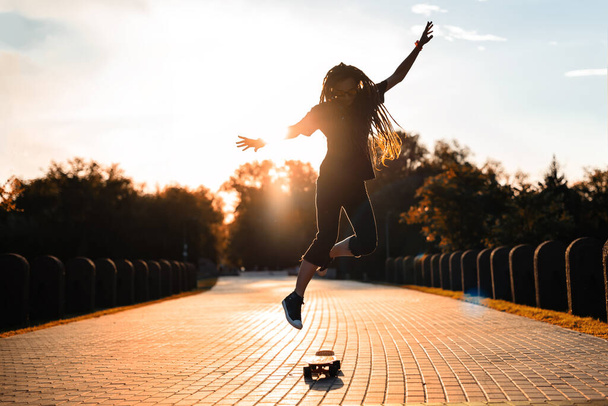 silhouette of a girl in gym shoes jumping on a skateboard in the setting sun in the park. hipster with dreadlocks and sunglasses learns tricks on a longboard - 写真・画像