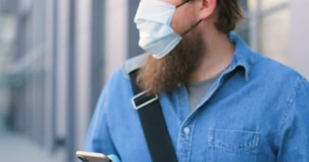 Close up of Caucasian man with beard and in medical mask texting message on mobile phone at street. Male pedestrian in respiratory protection tapping and scrolling on smartphone outdoors. - Imágenes, Vídeo