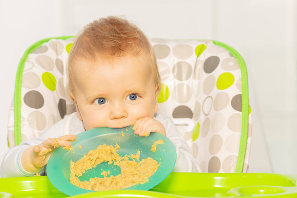 Toddler boy sit in high chair and play with porridge in plastic plate - Фото, зображення