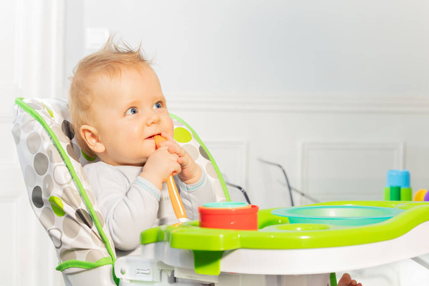Happy little toddler baby boy sit in highchair holding plastic spoon in mouse with thoughtful look - Φωτογραφία, εικόνα