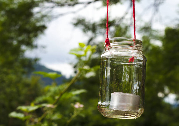 чайна світла свічка в склянці, що висить з дерева на червоній мотузці з розмитими рослинами на фоні
 - Фото, зображення