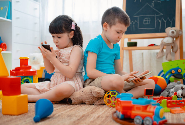 Fratelli bambini fratello e sorella, amici siedono sul pavimento della casa nella sala giochi per bambini con smartphone, distaccati dai giocattoli sparsi.
. - Foto, immagini