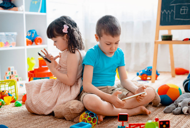 Siblings children brother and sister, friends sit on the floor of the house in the childrens play room with smartphones, detached from the scattered toys. - Zdjęcie, obraz