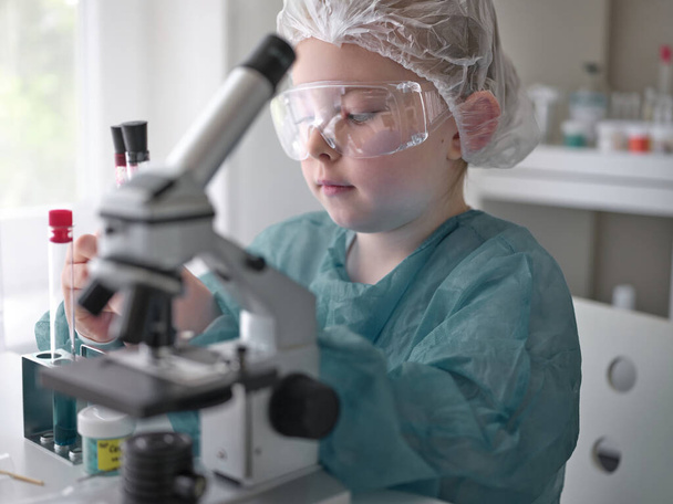 Милая маленькая девочка смотрит в микроскоп за его столом дома. Молодой ученый проводит эксперименты в своей лаборатории
.  - Фото, изображение