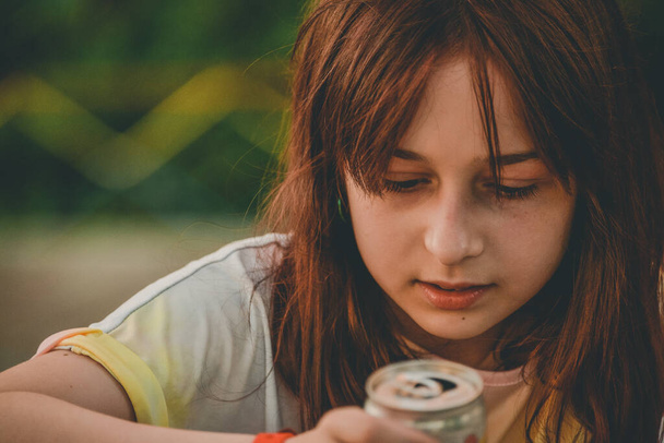 Młoda dziewczyna pije wodę. Nastolatka pije z żelazną puszką. Dziewięciolatka trzyma drinka w rękach. Nastolatka 9 lub 10 lat z brązowymi włosami. - Zdjęcie, obraz