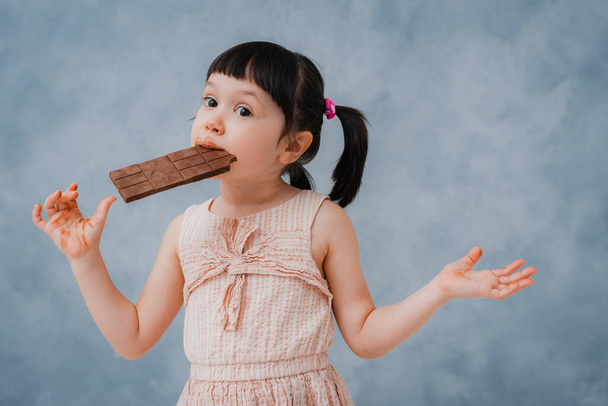 pequeña niña come chocolate y se lame el dedo sobre un fondo gris azul Studio. espacio libre
. - Foto, imagen