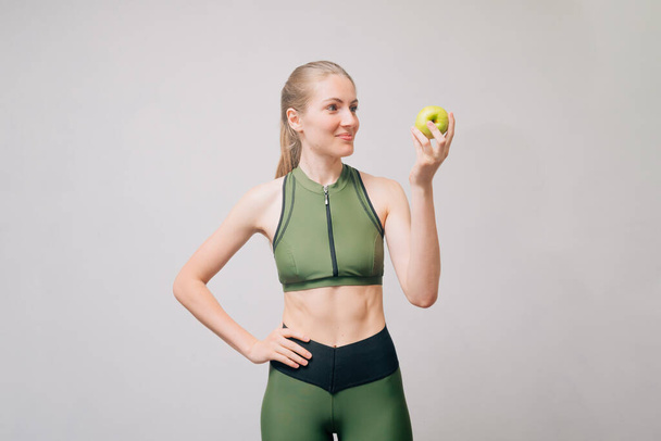 Sporty slim girl holding a green apple in her hands. Fitness model in a short theme and leggings. Happiness concept with smile slimming young woman. - Photo, Image