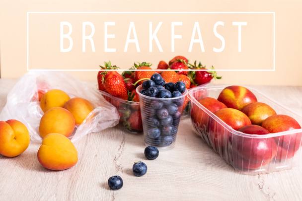 blueberries, strawberries, nectarines and peaches in plastic containers on wooden surface near breakfast lettering on beige - Photo, Image