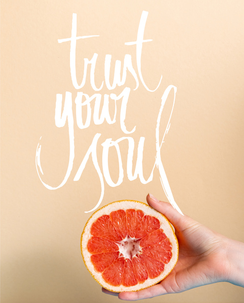 oříznutý pohled na ženskou ruku s šťavnatým grapefruitem zpola blízkým důvěře vaše duše naškrábaná na béžové  - Fotografie, Obrázek