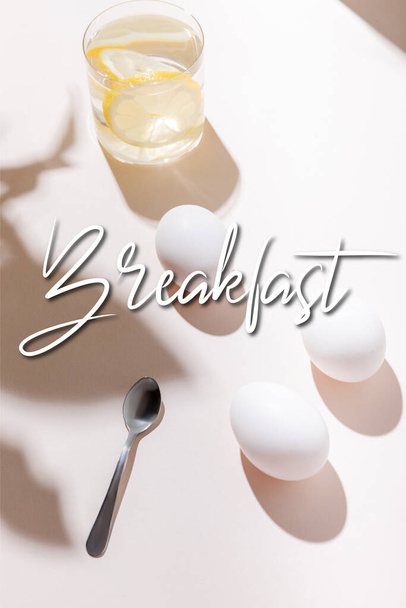 chicken eggs, teaspoon and glass of water with lemon on grey table with breakfast lettering - Photo, image