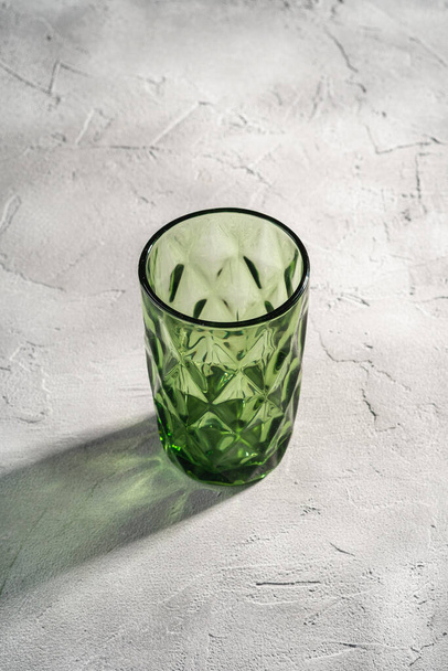 Green glass geometric cup with colorful shadow light rays on stone concrete background, angle view - Foto, imagen