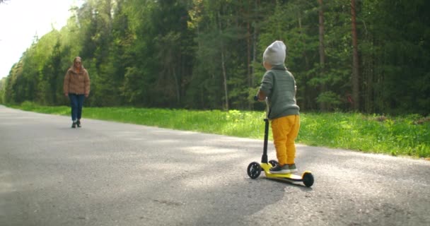 Chłopiec uczy się jeździć na skuterze i w zwolnionym tempie idzie spotkać się z matką na skuterze na drodze w parku z sosnami - Materiał filmowy, wideo