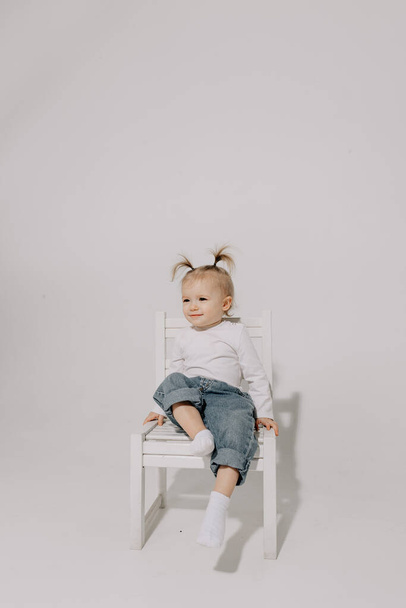 cute little girl with a ponytail hairstyle dressed in a white T-shirt and denim pants posing for a photo on a white background in a photo studio - Photo, image
