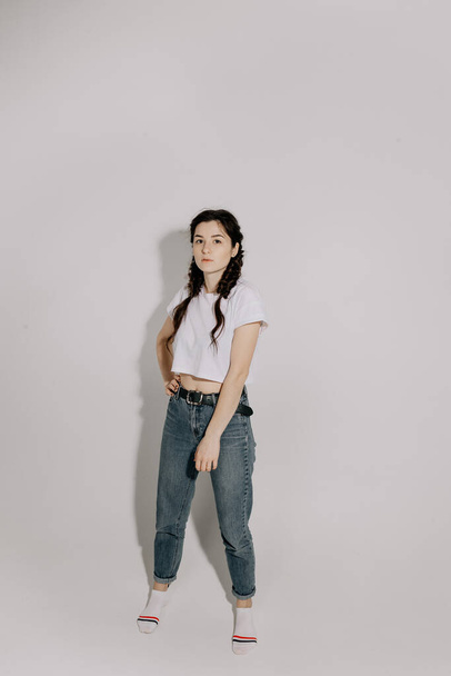 Young pretty woman with pigtail braid dressed in white t-shirt and jeans posing for photo on a white background in front of studio light - Photo, Image