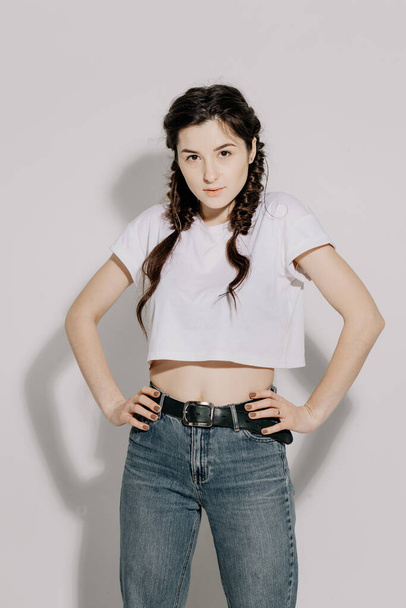 Young pretty woman with pigtail braid dressed in white t-shirt and jeans posing for photo on a white background in front of studio light - Фото, зображення
