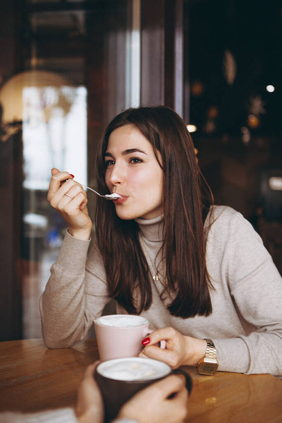 young pretty girl with a big pink mug of sweet coffee sits on a chair in her favorite cafe and drinks her daily morning coffee and talks with a girlfriend - Foto, imagen