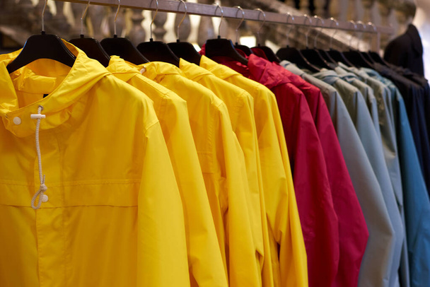 Capas de chuva coloridas brilhantes do outono penduradas em uma loja de moda. Closeup de multi-colorido amarelo, vermelho, cinza, impermeável impermeáveis pendurar em uma fileira em um cabide na loja de roupas no shopping
. - Foto, Imagem