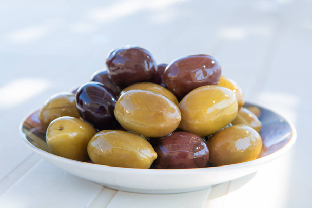 Aceitunas verdes y negras italianas en aceite de oliva de cerca servidas al aire libre sobre mesa blanca
 - Foto, Imagen
