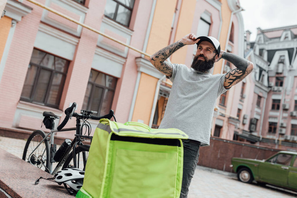 Always on time. Brutal stylish delivery man in cap looking aside, standing with backpack outdoors, ready to go after completing food delivery. Courier, delivery service concept - Foto, Imagem