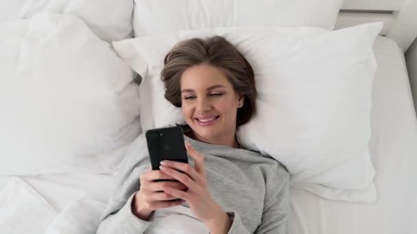 A happy smiling young woman is watching something good on her smartphone while lying in the white bed at home - Imágenes, Vídeo