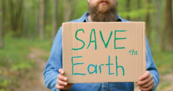 Primer plano del póster de cartón con palabras Save the Earth en manos del hombre caucásico. Hombre eco activista de pie en el bosque o parque en un día soleado con una sola protesta
. - Imágenes, Vídeo