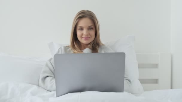 An attractive young woman is using her laptop computer lying in her white bed at home in the morning - Filmagem, Vídeo