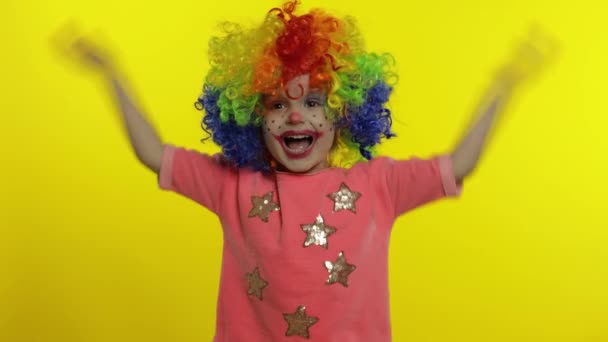 Malá holčička klaun v barevné paruce dělat hloupé obličeje, křičí, chytí ji za hlavu, mává rukama - Záběry, video
