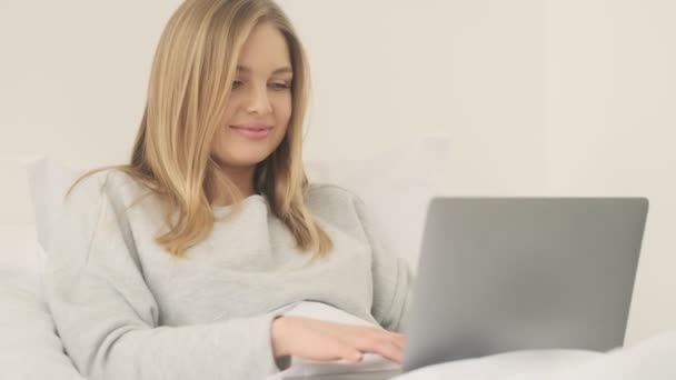 A lovely young woman is typing on her laptop computer in her white bed at home in the morning - Video