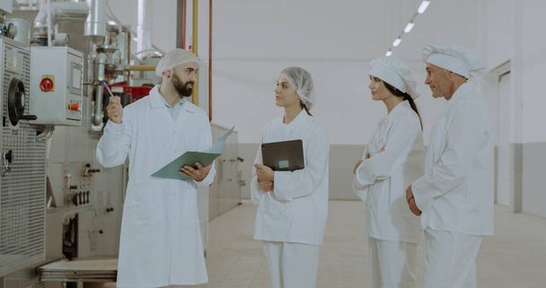 Food industry engineer man holding a tablet in hand and explaining the instructions about the machine to his workers they wearing special clothes for protection - Photo, Image