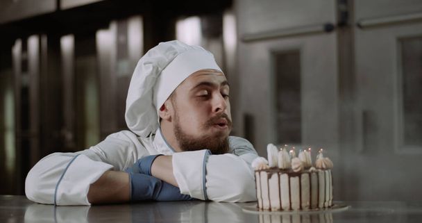 青い手袋を身に着けている若い美しい男と料理人の制服はケーキの上にろうそくを吹き消す - 写真・画像