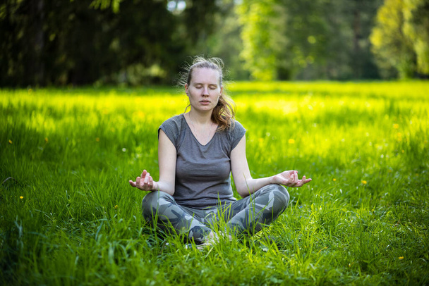Yoga en el parque. Mujer en posición de loto se sienta en la hierba verde en el parque. El sol ilumina la hierba en el fondo. La chica cerró los ojos.
. - Foto, Imagen