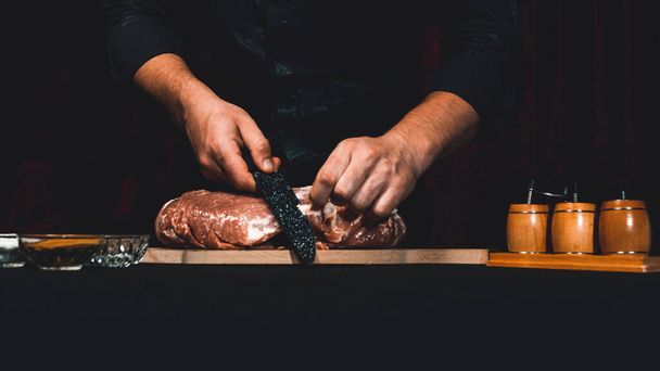 The chef of the restaurant in a dark uniform cuts the meat with a black kitchen knife. Preparation of pork marinated steak to order. Cutting off excess fat. - Zdjęcie, obraz