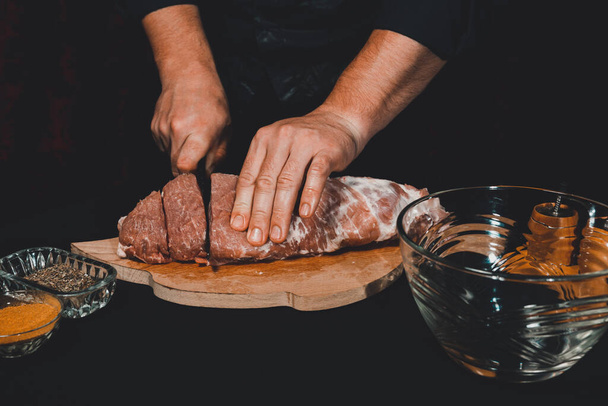 The chef of the restaurant in a dark uniform cuts the meat with a black kitchen knife. Preparation of pork marinated steak to order 2021. - Photo, Image