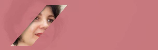 Il volto di una donna attraente di mezza età sbircia in un buco in carta rosa. Moda, bellezza, trucco, cosmetici, salone di bellezza, stile, cura personale, geometria. - Foto, immagini
