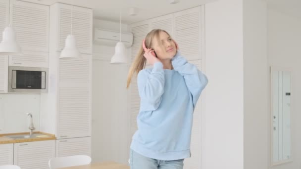 A smiling young blonde woman is enjoying listening the music using headphones in the living room at home - Кадри, відео